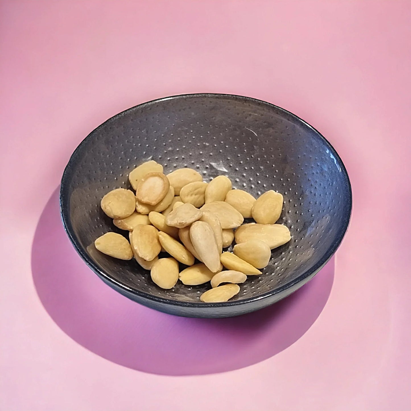 Mandorle pelate in ciotola nera su sfondo rosa, ingrediente per dolci e pasticceria