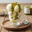 Gelato artigianale alla crema con salsa al pistacchio e pistacchi interi su vassoio di legno.